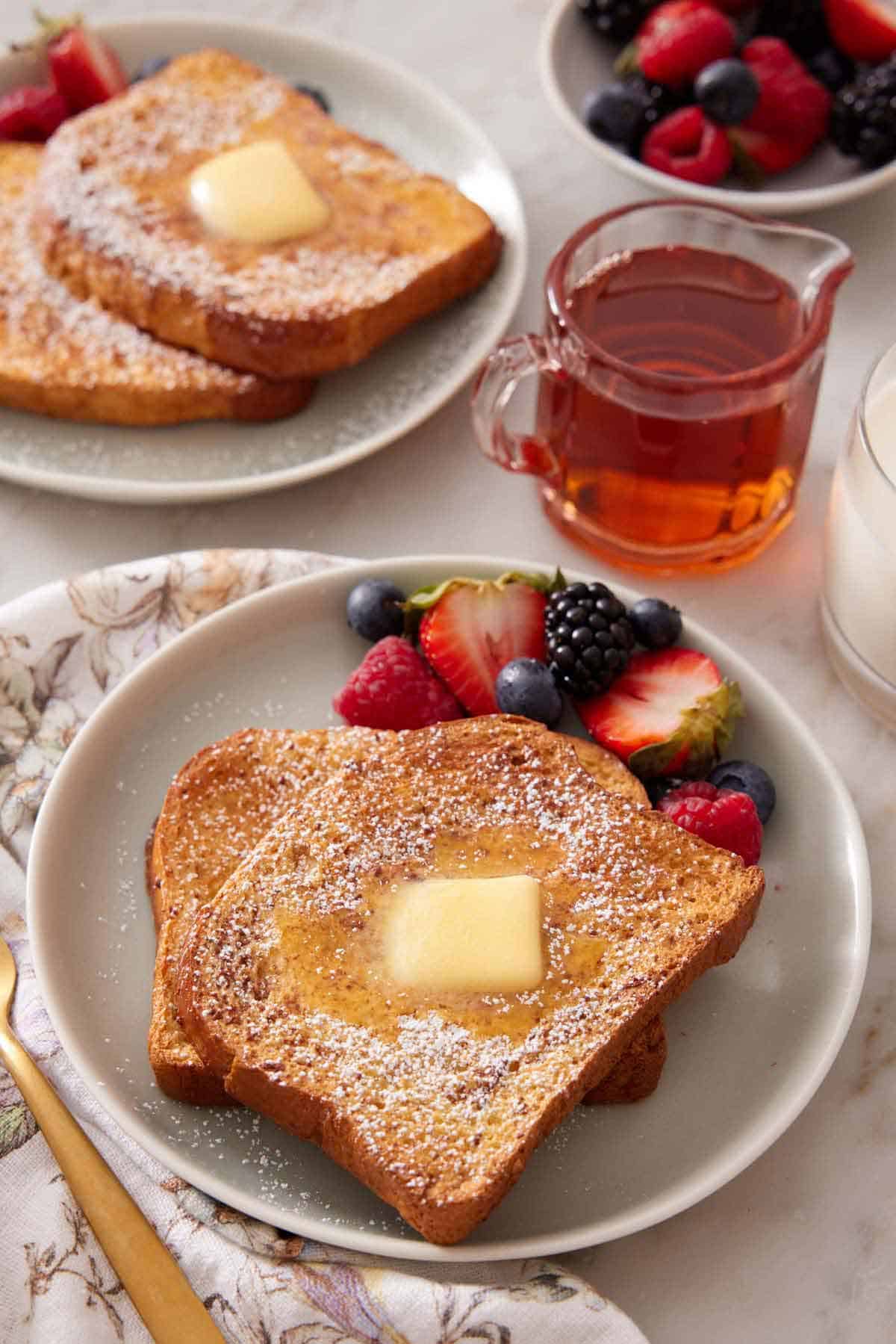 A plate of air fryer french toast with powdered sugar, butter and syrup on top with berries on the side. A small vessel of syrup and another plated serving in the background.
