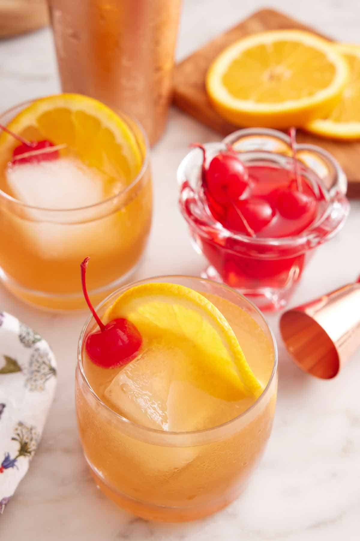 Two glasses of Amaretto Sour garnished with orange slices and maraschino cherries with a jar of more cherries and sliced orange on a cutting board in the back.