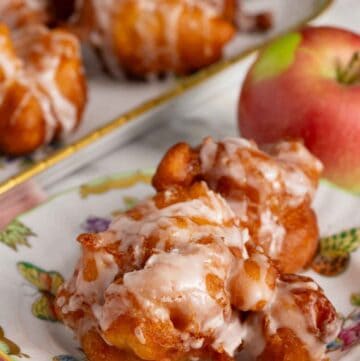 A plate with apple fritters topped with glaze with an apple and platter of more fritters in the background.