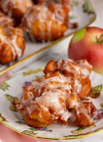 A plate with apple fritters topped with glaze with an apple and platter of more fritters in the background.