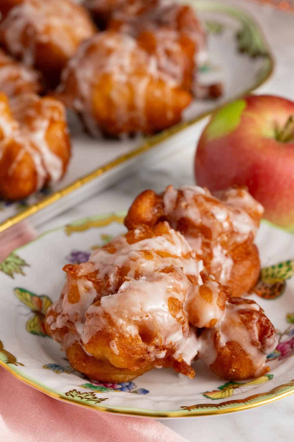 A plate with apple fritters topped with glaze with an apple and platter of more fritters in the background.