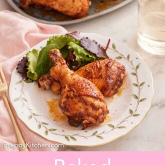 Pinterest graphic of a plate with two baked chicken legs with some salad with a platter in the background along with wine.