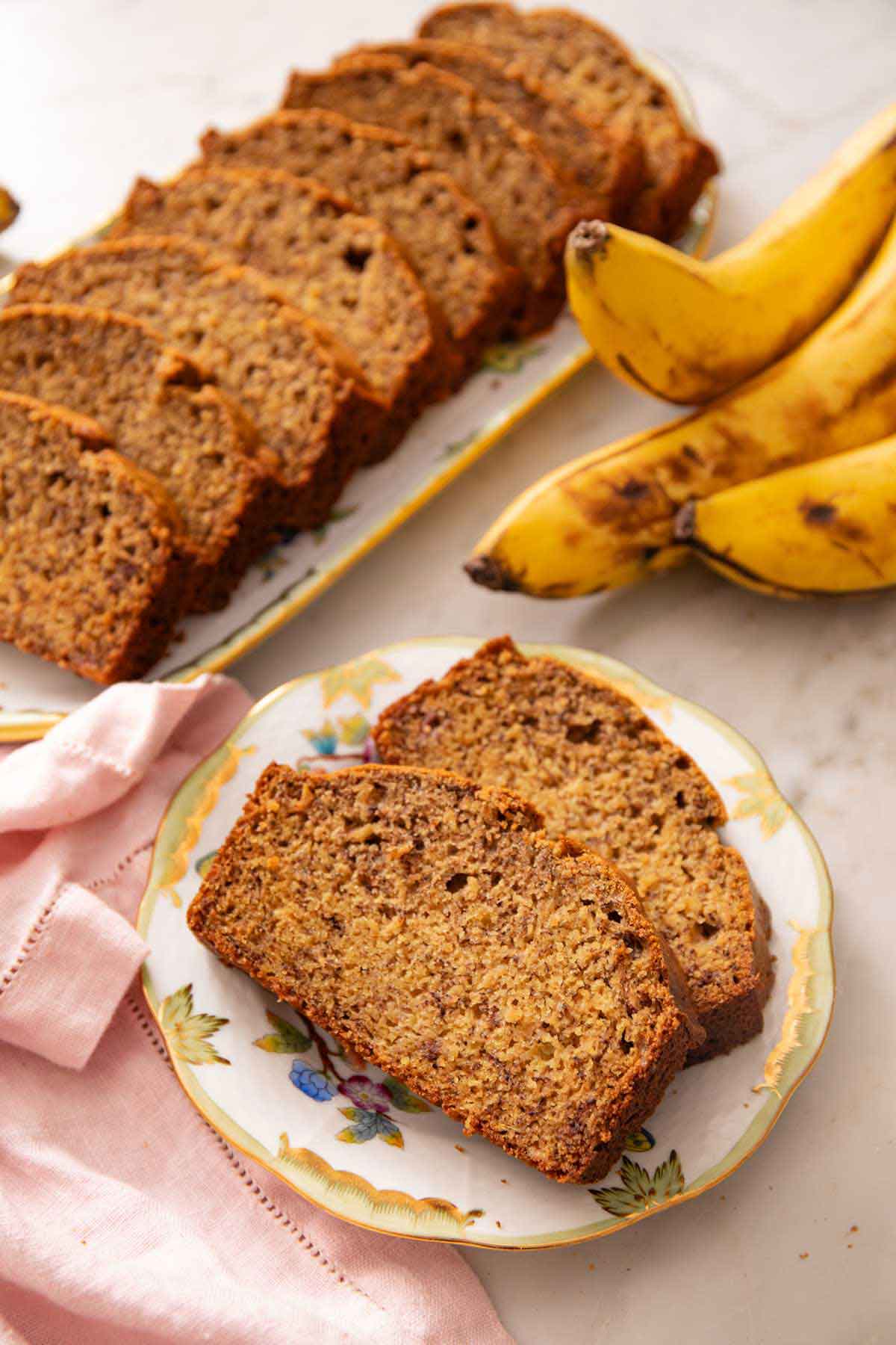 A plate with two slices of banana bread with a platter in the background with the rest of the sliced loaf. Bananas off to the side.