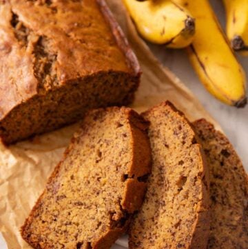 A loaf of banana bread with three slices in front. Bananas in the background.