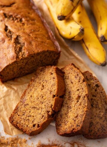 A loaf of banana bread with three slices in front. Bananas in the background.