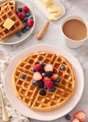 A plate with a Belgian waffle topped with berries. Coffee, butter, and more waffles in the background.