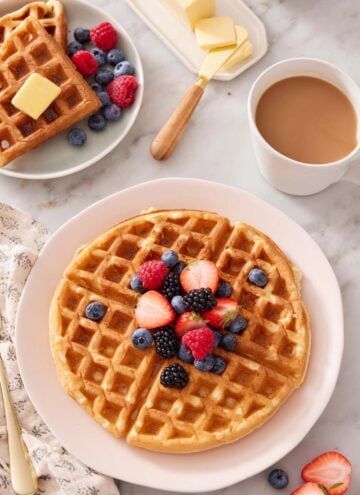 A plate with a Belgian waffle topped with berries. Coffee, butter, and more waffles in the background.