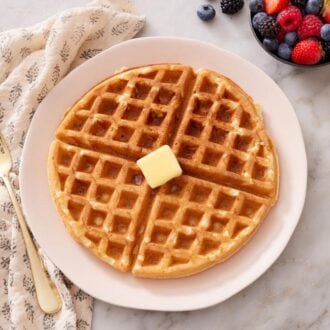 Overhead view of a Belgian waffle topped with a knob of butter. Fork and bowl of berries to the side.