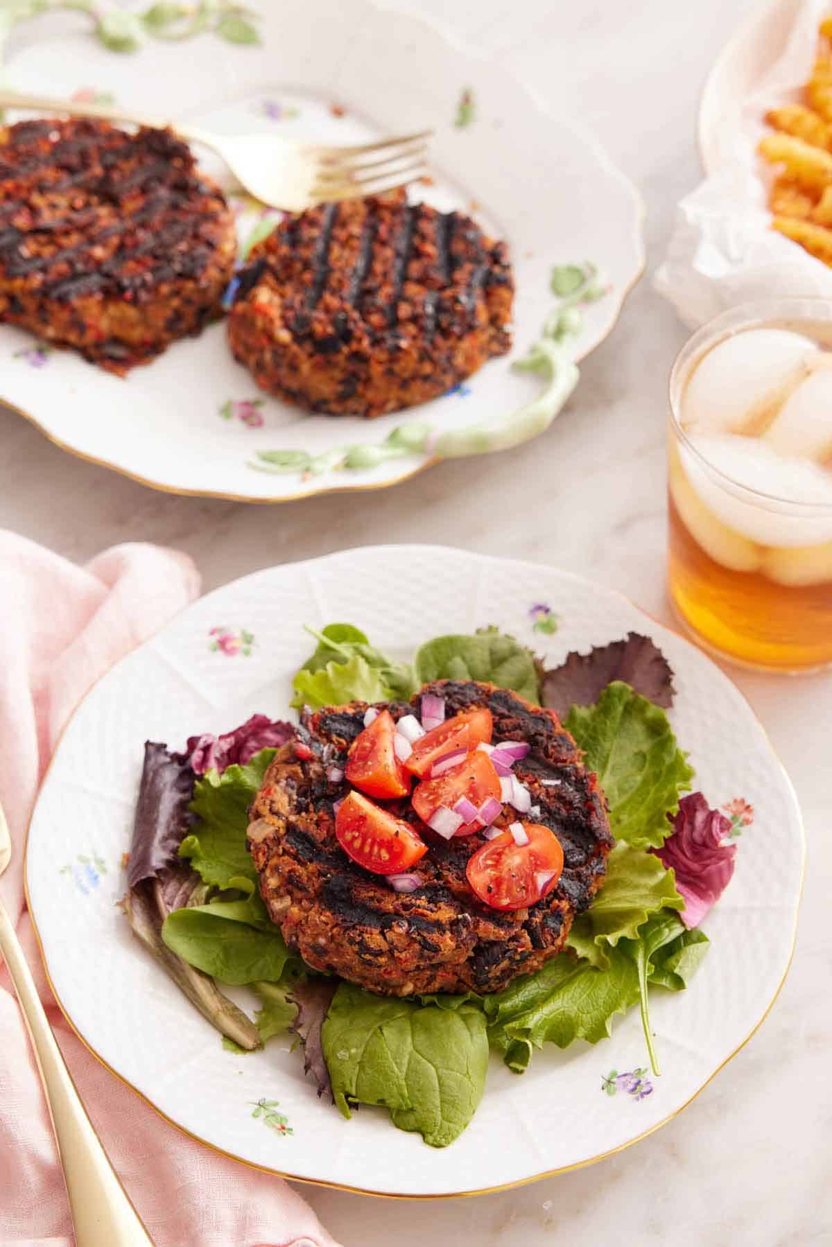 A plate with a black bean burger patty topped with tomatoes and red onions over a bed of lettuce. More patties in the background.