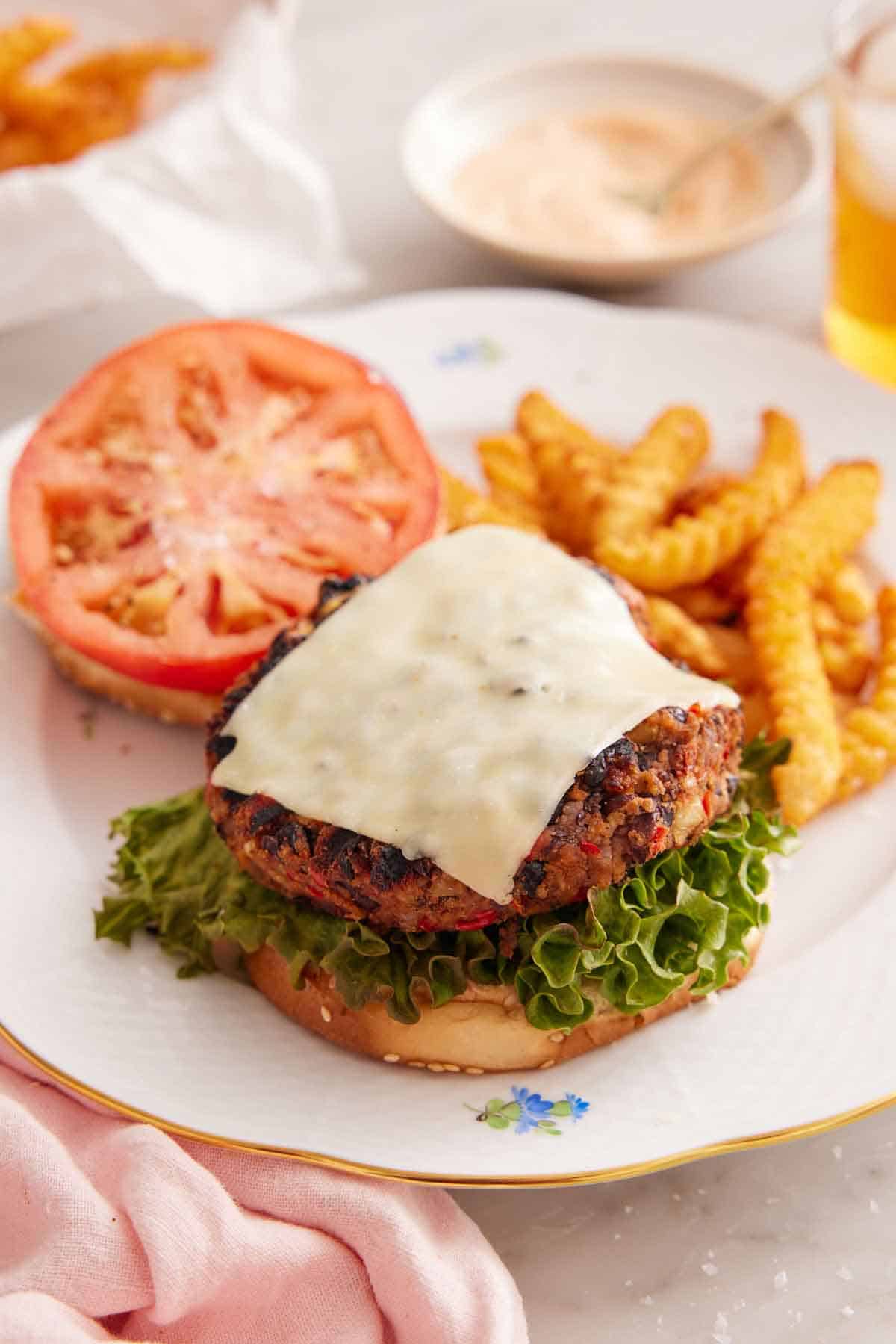 A plate with a black bean burger with a slice of melted cheese on top with the burger bun with a tomato slice in the background.