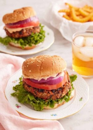A plate with a black bean burger with a drink in the back along with a second burger and bowl of fries.