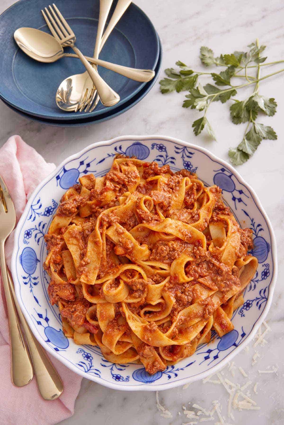 Overhead view of a bowl of bolognese. A stack of plates, utensil, and parsley in the background.