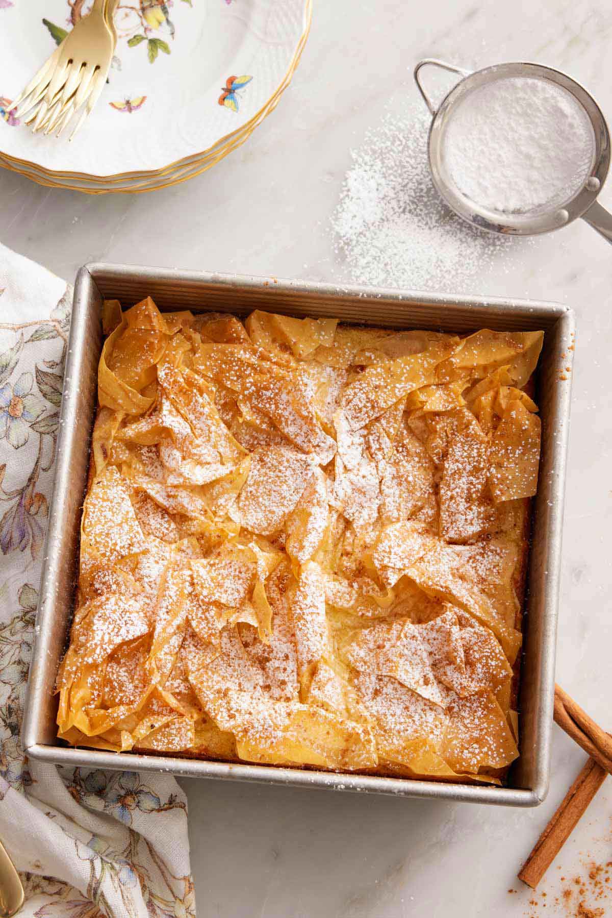 Overhead view of a square pan of Bougatsa with a sifter of powdered sugar to the side.