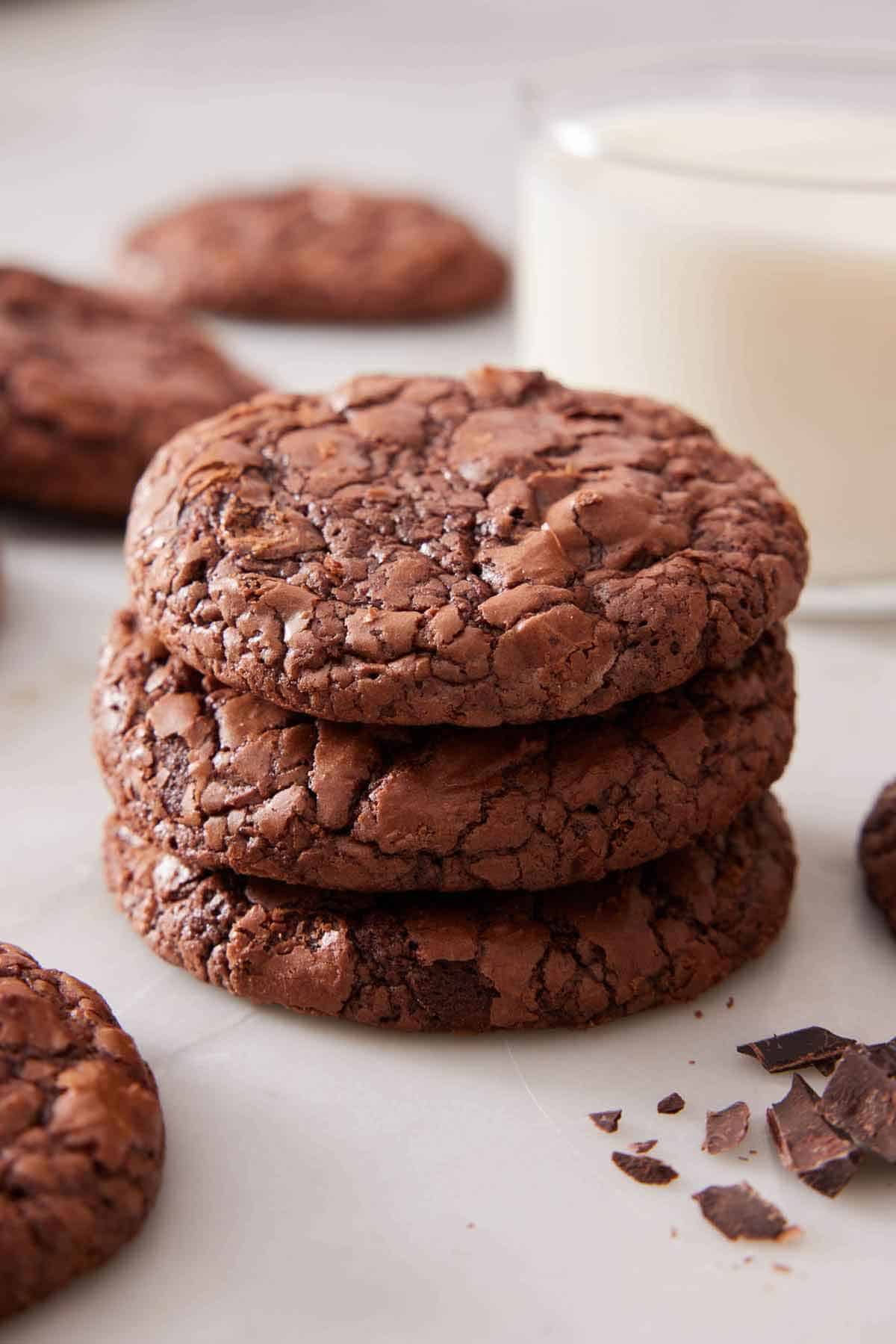 A stack of three brownie cookies with a cup of milk in the background and more cookies and chopped chocolate scattered around.