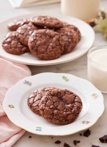 A plate with two brownie cookies with a cup of milk and platter of more cookies in the background.
