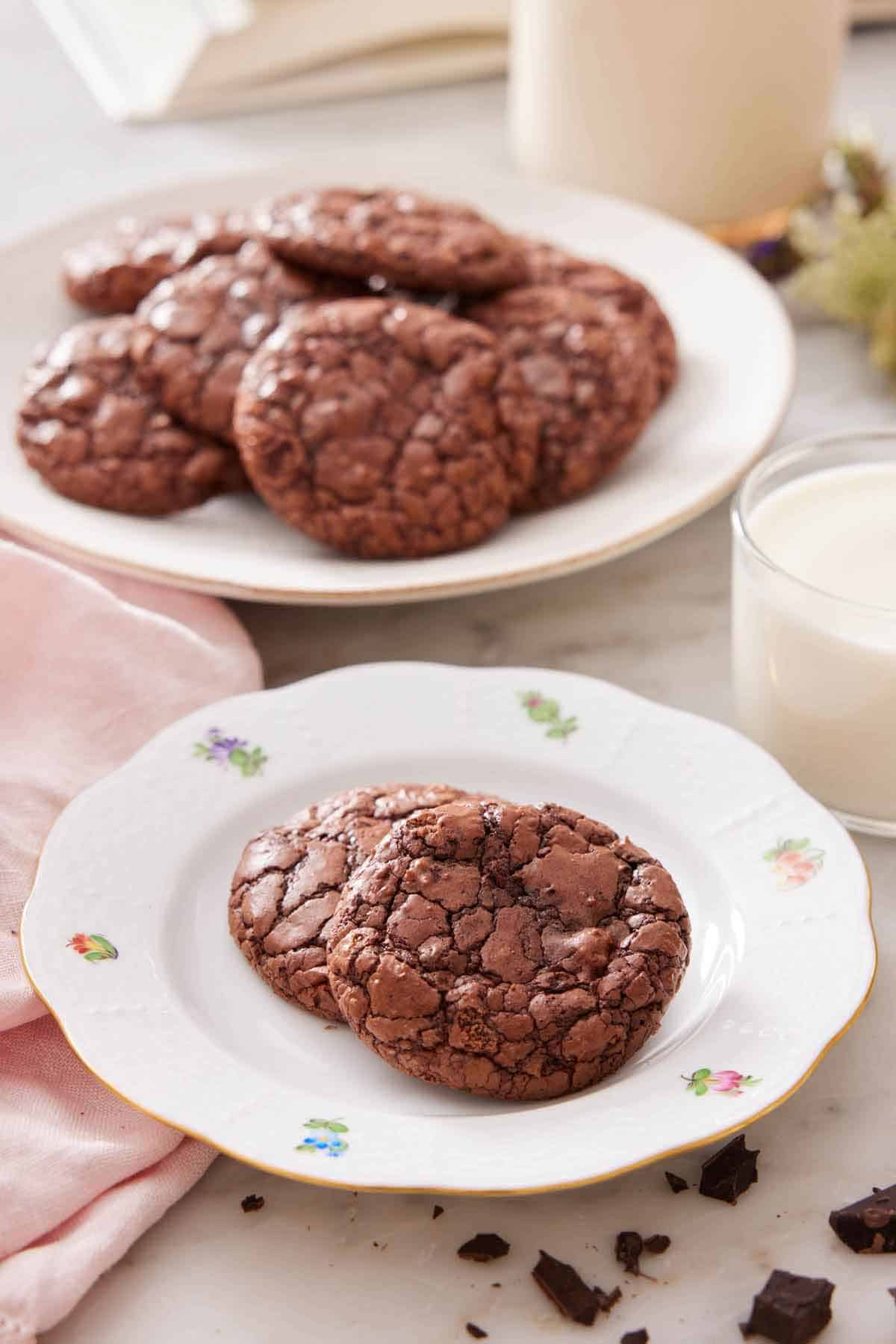 A plate with two brownie cookies with a cup of milk and platter of more cookies in the background.