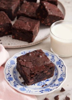 A plate with a piece of brownie with a cup of milk and platter of brownies in the background.