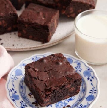 A plate with a piece of brownie with a cup of milk and platter of brownies in the background.
