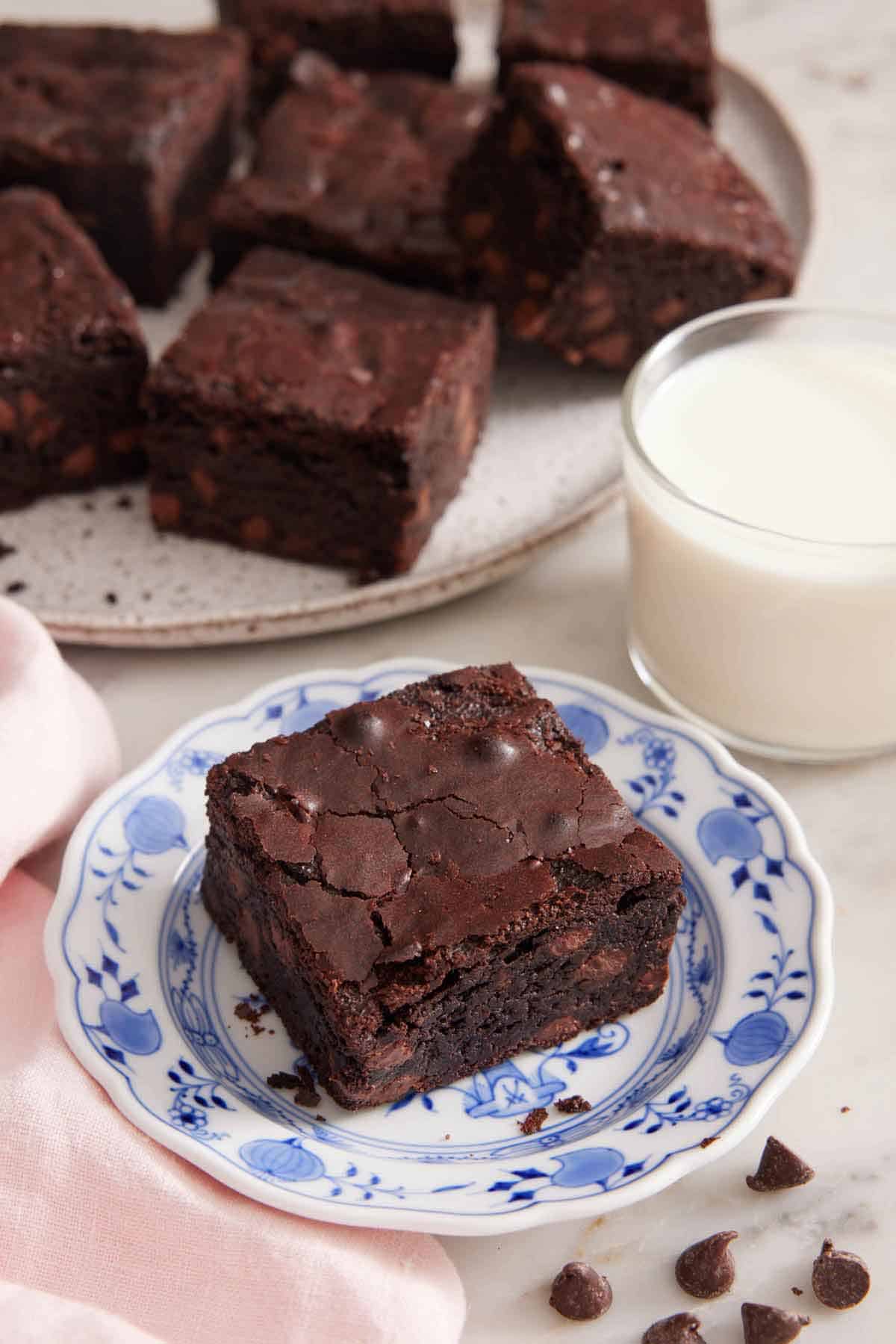 A plate with a piece of brownie with a cup of milk and platter of brownies in the background.