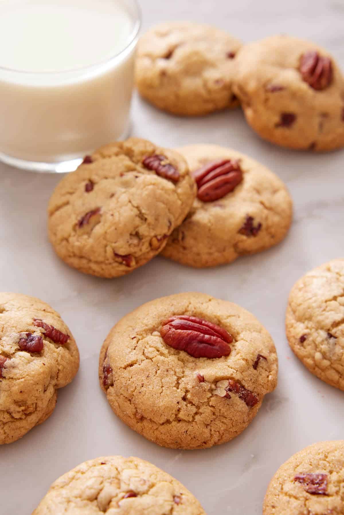 Multiple butter pecan cookies scattered on a marble counter with a glass of milk.