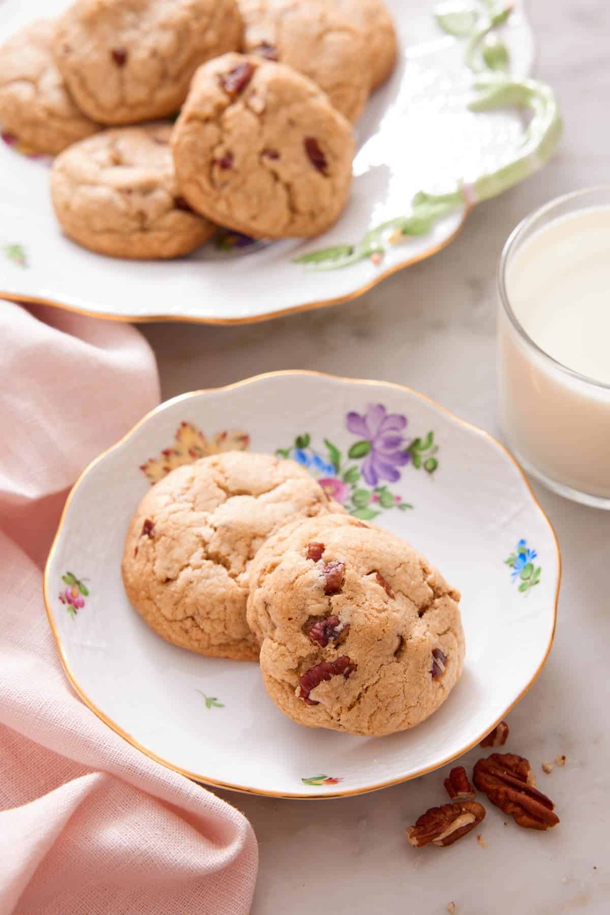 Two butter pecan cookies on a plate with a glass of milk and platter in the background.