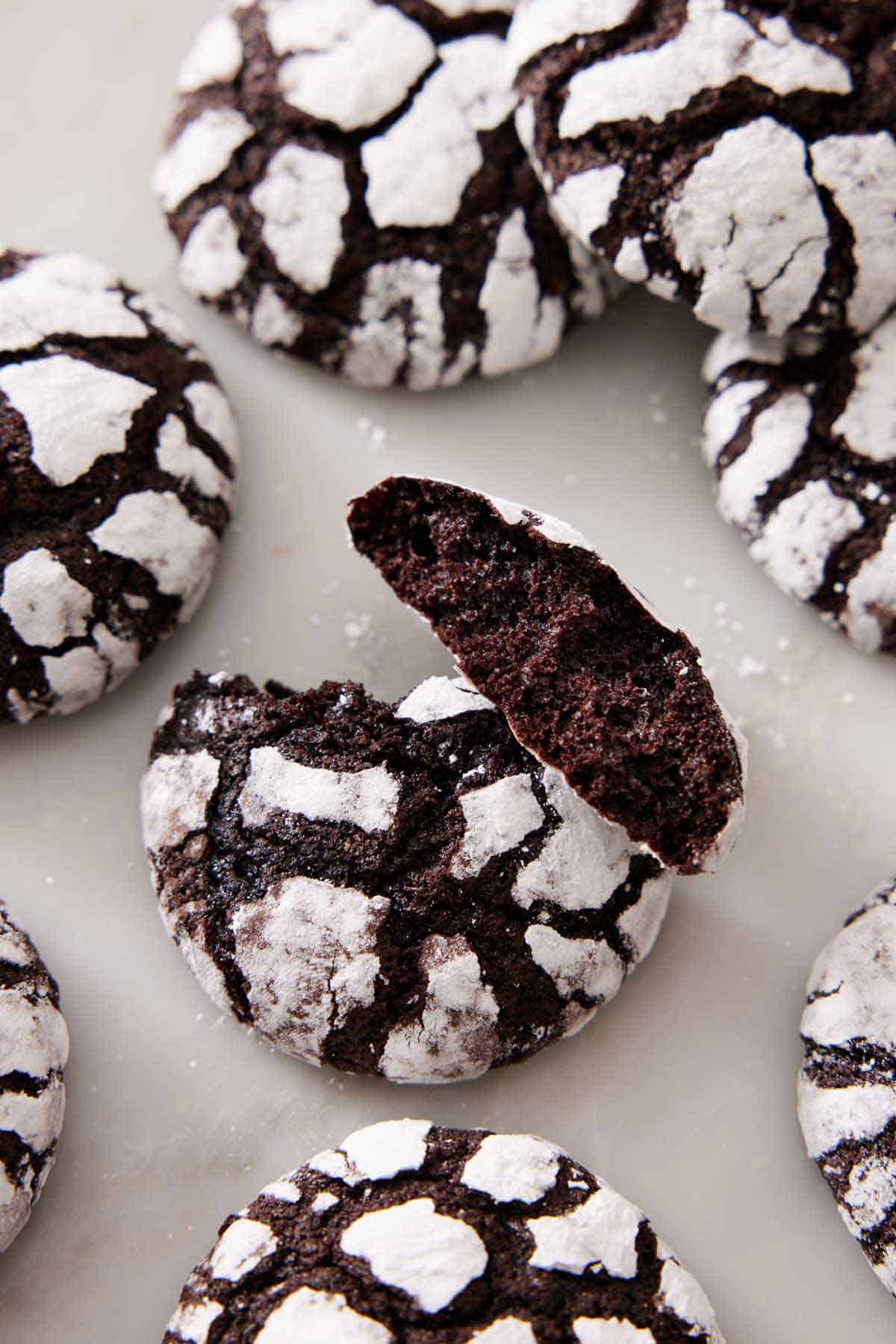 Chocolate crinkle cookies scattered on a marble surface with the one in the middle broke in half, showing the interior.