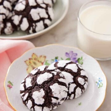 A plate with two chocolate crinkle cookies with a glass of milk and platter of more cookies in the background.