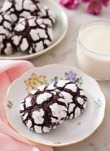 A plate with two chocolate crinkle cookies with a glass of milk and platter of more cookies in the background.