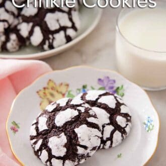 Pinterest graphic of a plate with two chocolate crinkle cookies with a glass of milk and platter of more cookies in the background.