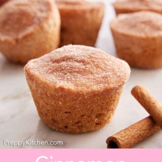 Pinterest graphic of a cinnamon muffin on a marble surface with more muffins in the back and some cinnamon sticks on the side.