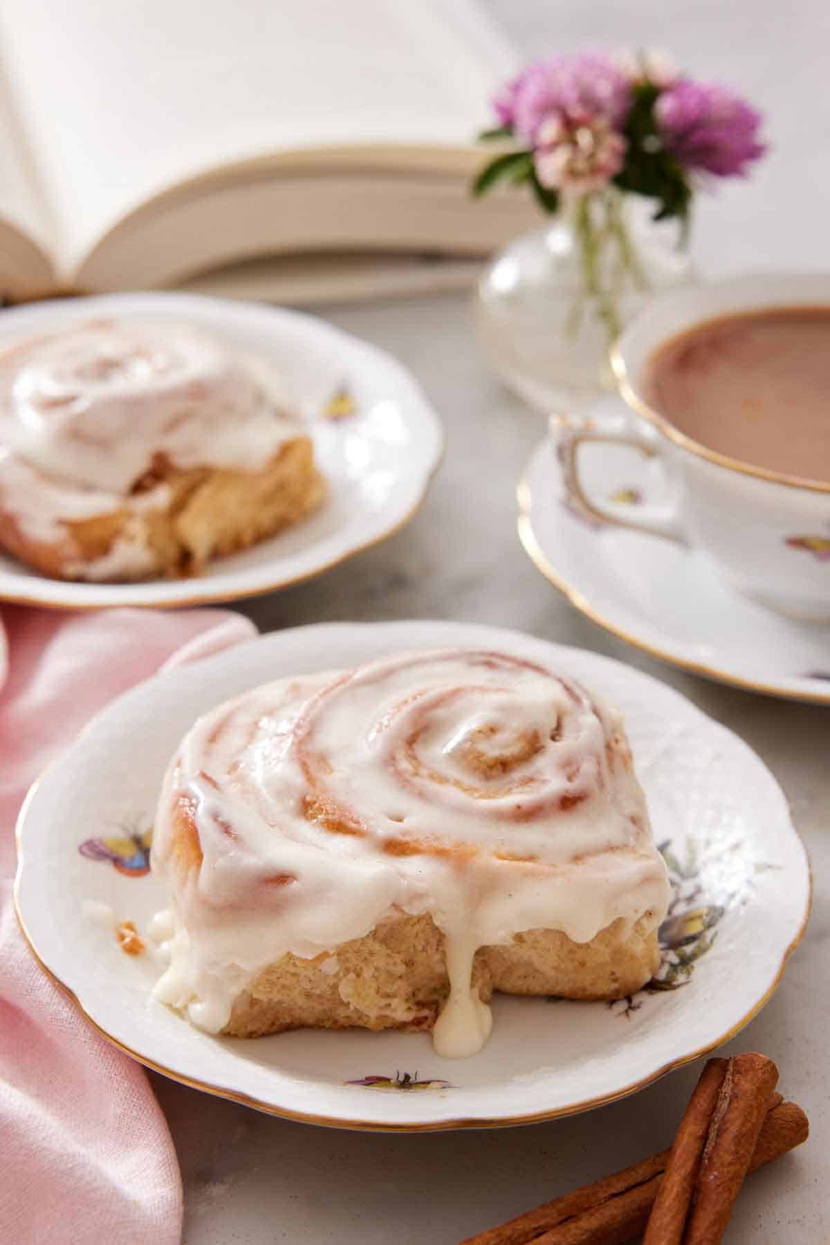 Two plates with cinnamon rolls and a cup of coffee in the background with some flowers and a book.