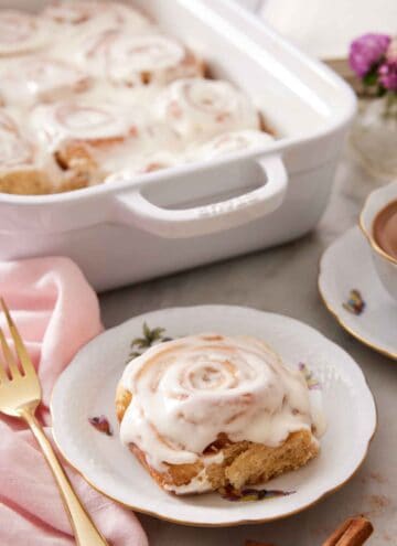 A plate with a cinnamon roll on a plate with a baking dish with more cinnamon rolls in the background.