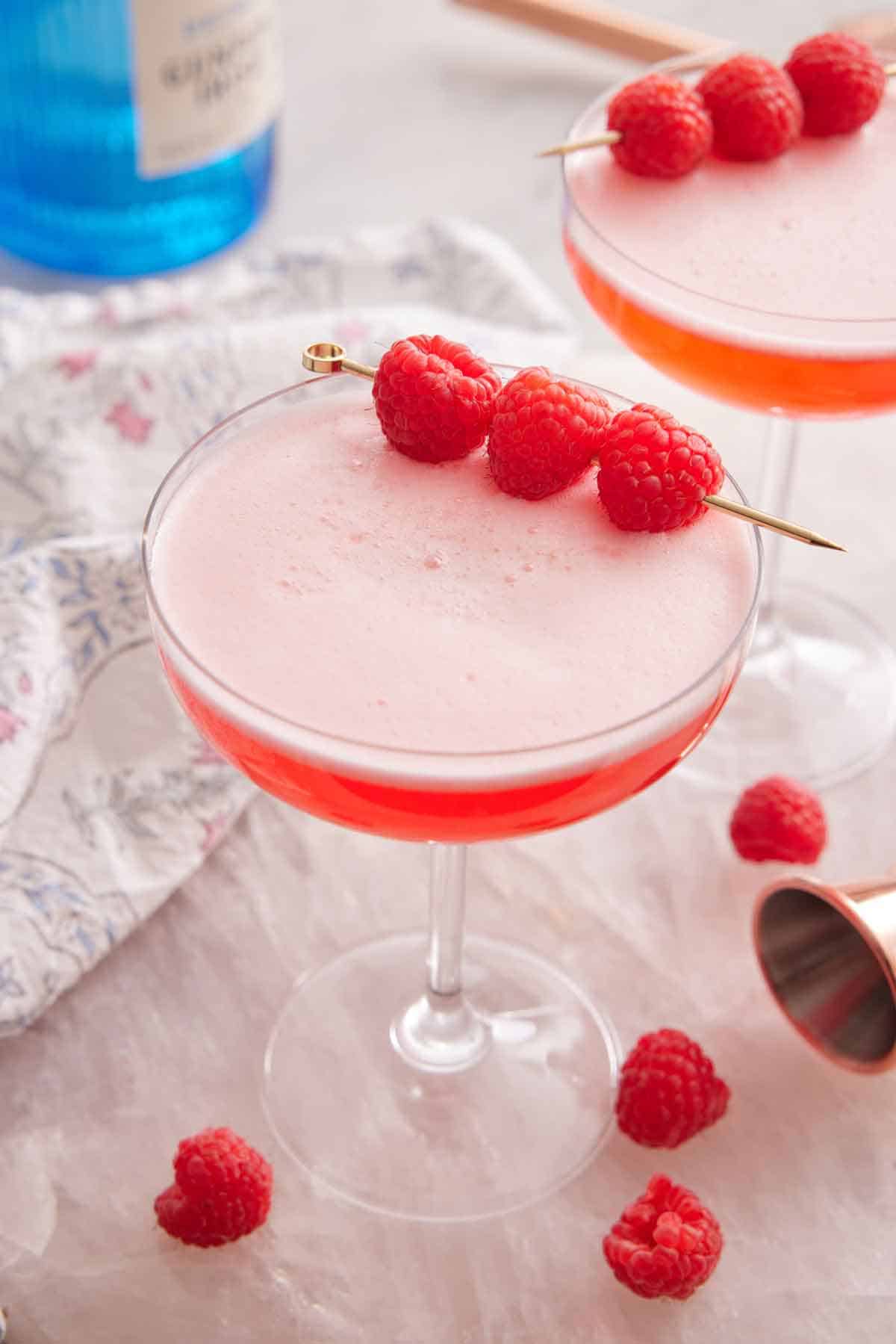 a slightly overhead view of a glass of Clover Club Cocktail with a second one in the background with raspberries scattered on the counter and as a garnish.