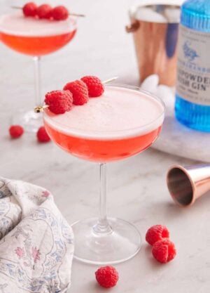 A glass of Clover Club Cocktail with raspberries scattered around and as a garnish. A bottle of gin and second cocktail in the background.