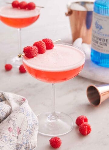 A glass of Clover Club Cocktail with raspberries scattered around and as a garnish. A bottle of gin and second cocktail in the background.