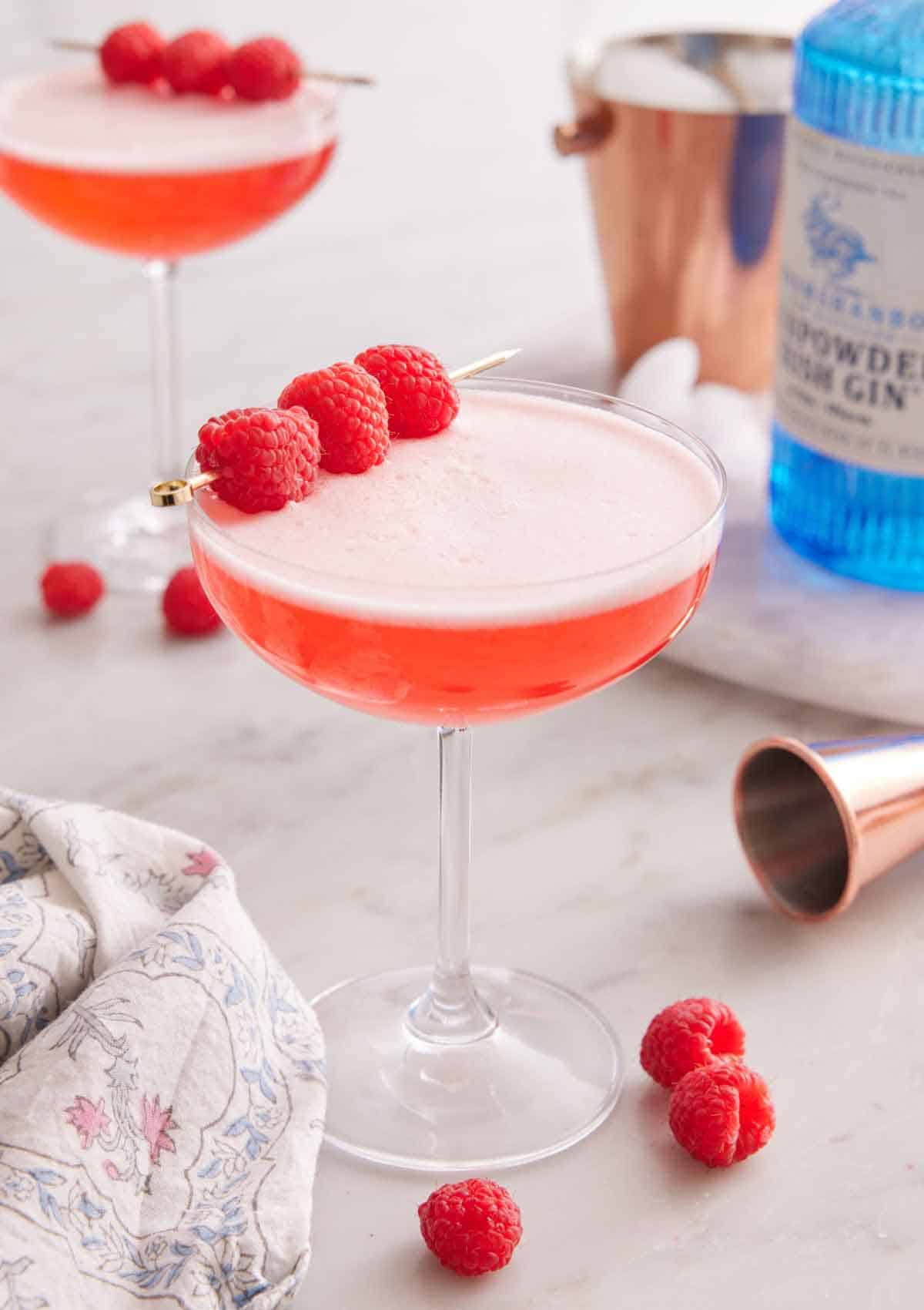 A glass of Clover Club Cocktail with raspberries scattered around and as a garnish. A bottle of gin and second cocktail in the background.