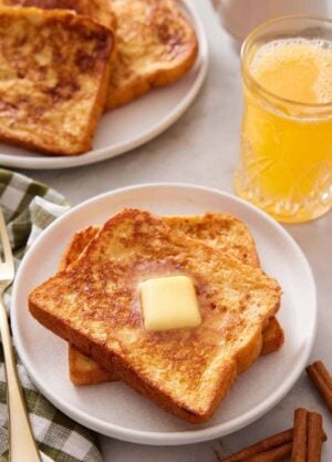 A plate with two pieces of french toast with a knob of butter on top. Orange juice and more french toast in the background.