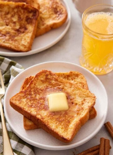 A plate with two pieces of french toast with a knob of butter on top. Orange juice and more french toast in the background.