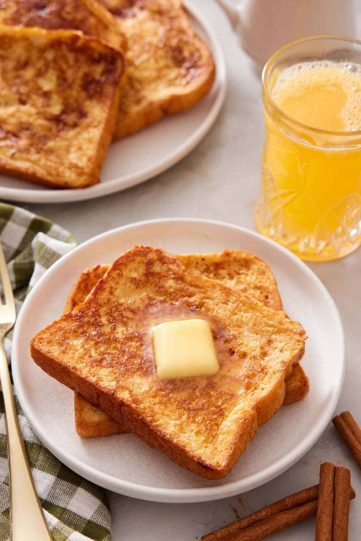 A plate with two pieces of french toast with a knob of butter on top. Orange juice and more french toast in the background.