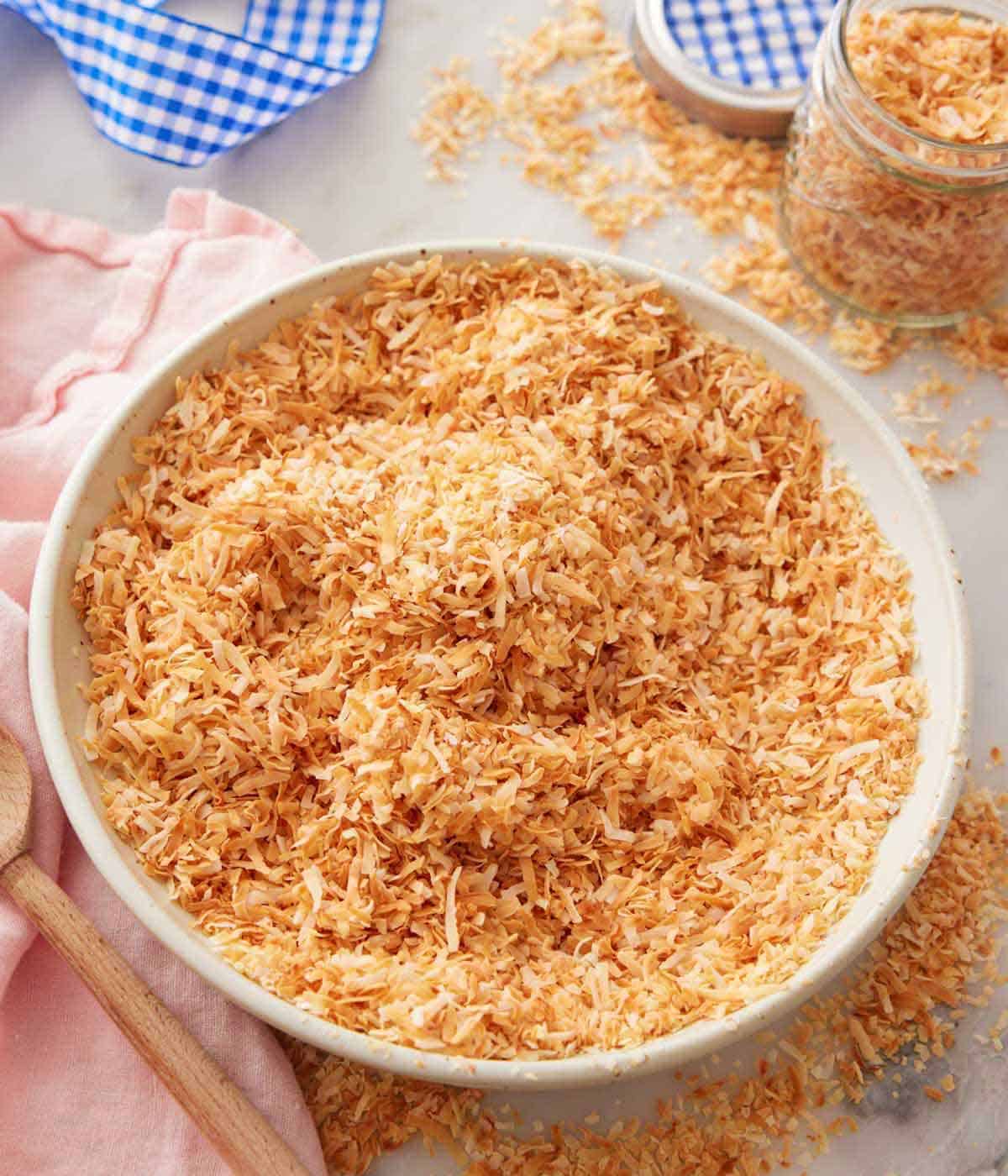 A large bowl of toasted coconut with a jar of to the side with more coconut scattered around.
