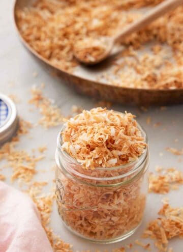 A jar of toasted coconut with a skillet in the background with more coconut.