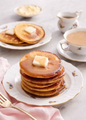 A stack of Johnny cakes with a cup of coffee and another plate with 3 Johnny cakes in the background along with a bowl of butter and syrup.