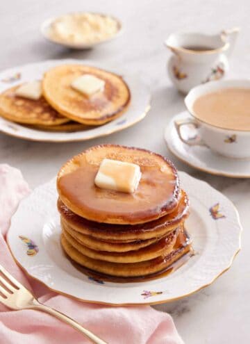 A stack of Johnny cakes with a cup of coffee and another plate with 3 Johnny cakes in the background along with a bowl of butter and syrup.