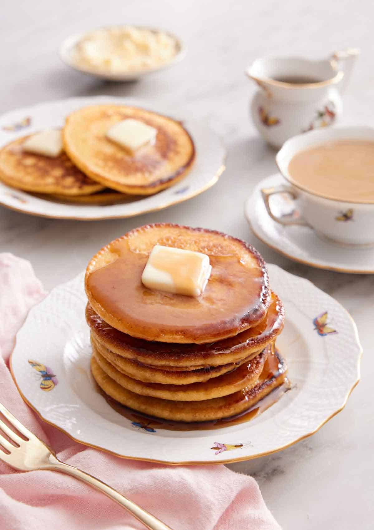 A stack of Johnny cakes with a cup of coffee and another plate with 3 Johnny cakes in the background along with a bowl of butter and syrup.