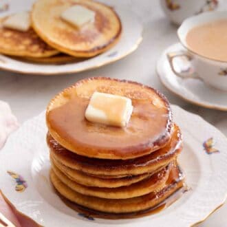 Pinterest graphic of a stack of Johnny cakes with a cup of coffee and another plate of Johnny cakes in the background along with a bowl of butter and syrup.