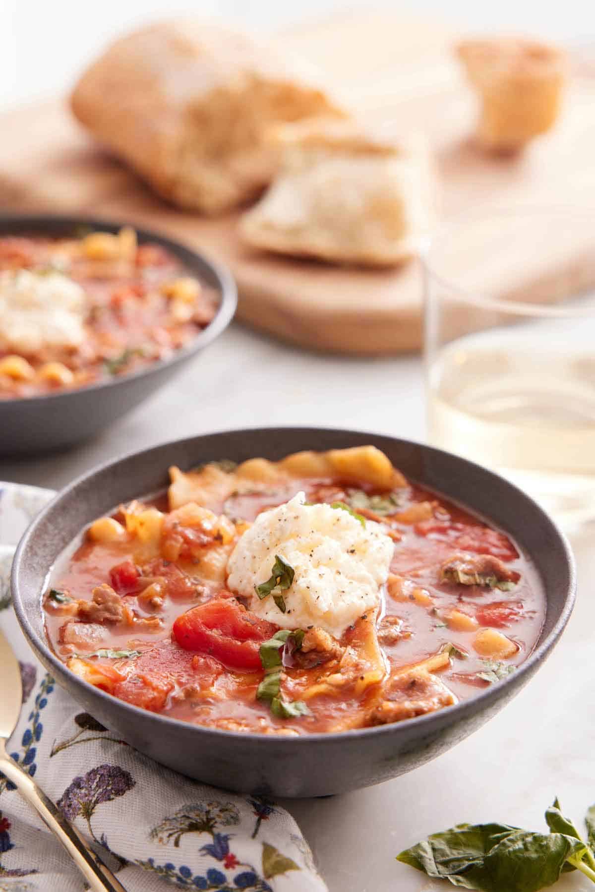 A bowl of lasagna soup with a glass of wine and torn bread in the background.