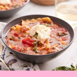 Pinterest graphic of a bowl of lasagna soup with a dollop of cheese in the middle and some torn bread, wine, and a second bowl of bowl in the background.