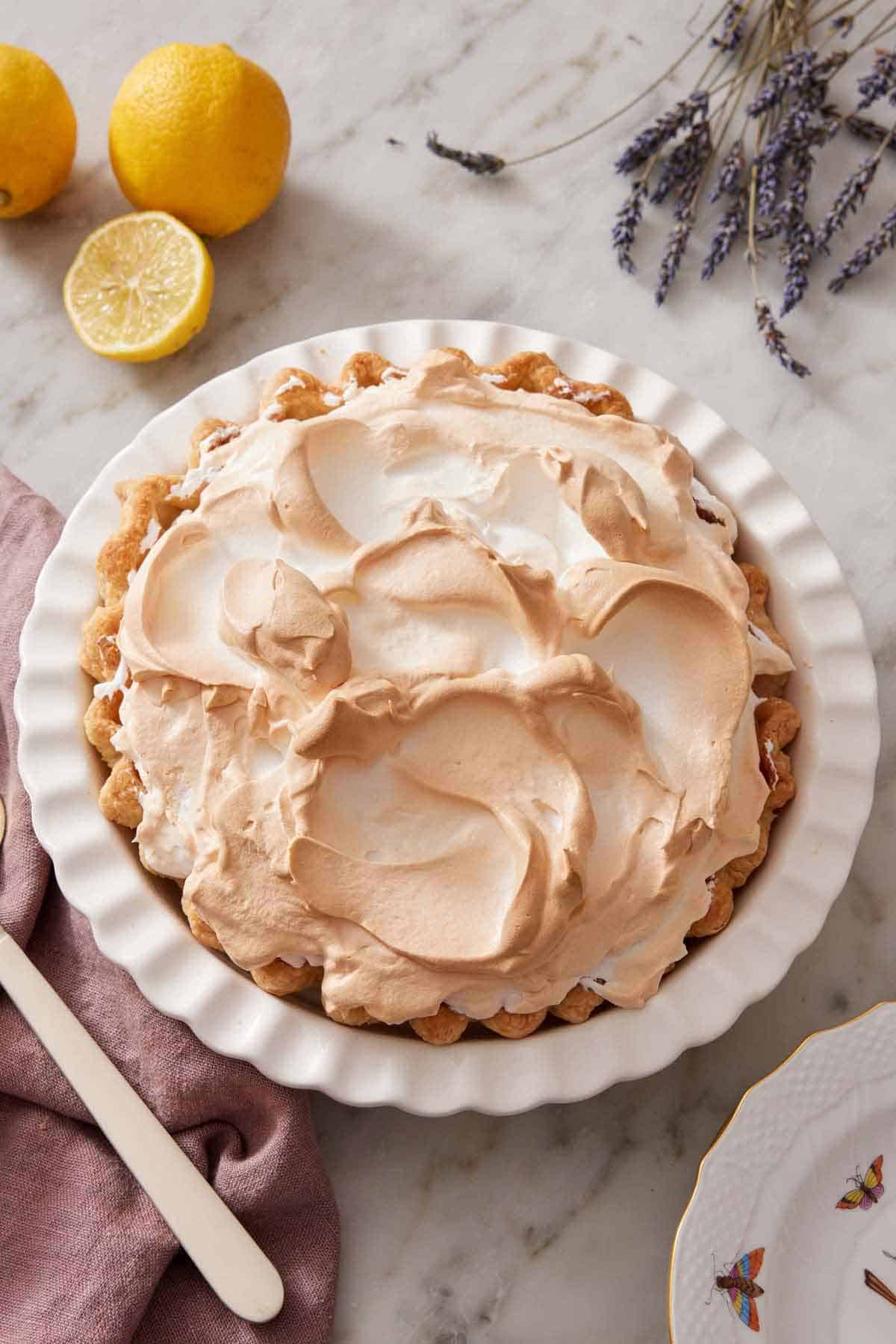 A whole lemon meringue pie in a white baking dish with some lemons and lavender to the side.