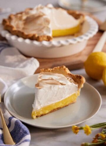 A slice of lemon meringue pie on a plate with the rest of the pie in the background.