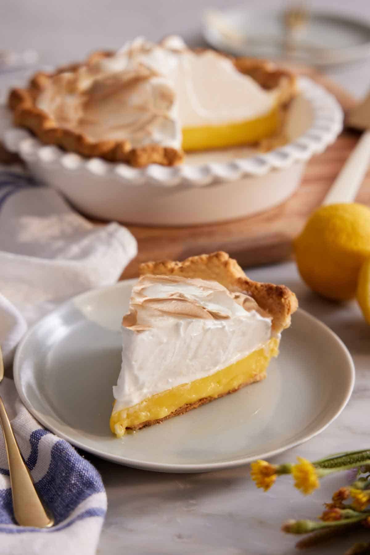 A slice of lemon meringue pie on a plate with the rest of the pie in the background.